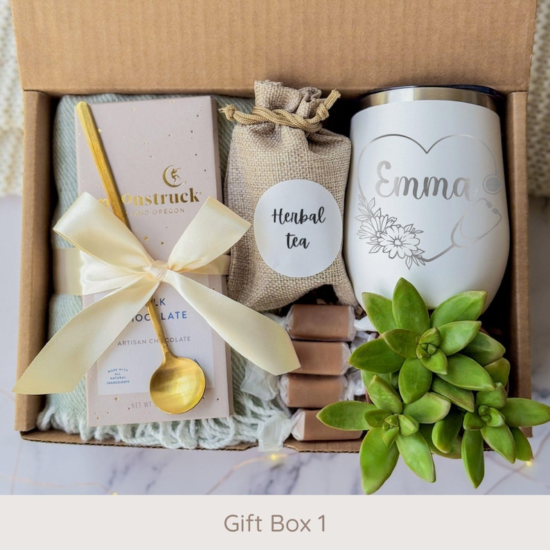 May include: A gift box filled with a variety of items. Includes a white tumbler with the name "Emma" and a floral design, a bar of chocolate, a gold spoon, herbal tea, and a succulent plant. The box is tied with a cream-colored ribbon. The text "Gift Box 1" is at the bottom.
