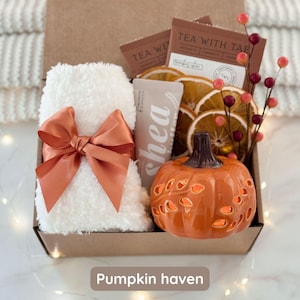 May include: A gift box with a white fluffy towel tied with an orange bow, a small orange ceramic pumpkin, tea packets, dried orange slices, and a shea butter tube. The box is labeled "Pumpkin haven".