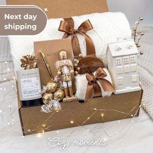 May include: A gift box containing a white knit blanket tied with a brown ribbon, a notebook, a gold pen, a nutcracker figurine, three gold-wrapped chocolates, a tea canister labeled Tea With Tae, and a white ceramic house-shaped candle holder.
