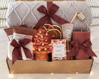 May include: A gift box containing a light gray textured throw blanket with a dark red ribbon, a ceramic light-up Christmas tree, dried orange slices, a Tea With Tae packet, a tube of hand cream, a maple soap bar, cinnamon sticks, and a red candle.
