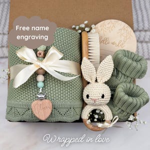 May include: A sage green knitted baby blanket with a cream satin ribbon, a wooden pacifier clip with the name Oliver, a wooden hairbrush, a crochet bunny rattle, and sage green knitted booties inside a gift box with a hello world wooden disc.