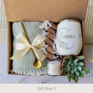 May include: A gift box filled with a light green blanket tied with a cream ribbon, a gold spoon, wrapped candies, a white tumbler with a floral design and the name "Emma", a small candle, and a succulent plant.