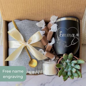 May include: A gift box with a gray blanket tied with a gold ribbon, a black tumbler with "Emma" and a stethoscope design, wrapped candies, a small candle, and a succulent plant. The box includes the text "Free name engraving".