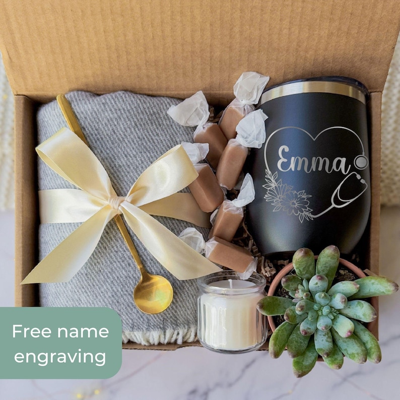 May include: A gift box with a gray blanket tied with a gold ribbon, a black tumbler with "Emma" and a stethoscope design, wrapped candies, a small candle, and a succulent plant. The box includes the text "Free name engraving".