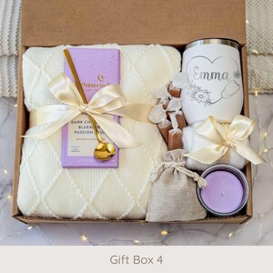 May include: A gift box filled with a cream-colored blanket, a purple chocolate bar, a gold spoon, a white tumbler with a heart design, candies, a small bag, and a lavender candle. The box is tied with a cream ribbon. The text "Gift Box 4" is at the bottom.