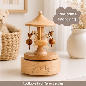 May include: A light brown wooden carousel music box featuring three carved horses suspended from a central canopy. The base of the carousel is engraved with the name Emily in a cursive script. The text Free name engraving is visible.