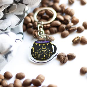 May include: A black and gold coffee mug keychain with a purple liquid inside and a crescent moon and eye design. The keychain is made of clear acrylic and is surrounded by coffee beans.