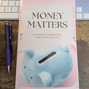 May include: A pink and white book titled "Money Matters: A Savings Workbook for Every Budget" with a blue piggy bank illustration on the cover.