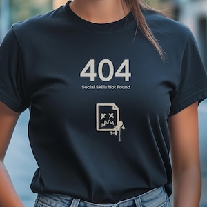Può includere: Una maglietta nera con un design grafico bianco che dice "404 Social Skills Not Found". La grafica include anche un'immagine stilizzata di un documento con un viso triste.