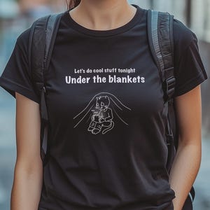 May include: Black t-shirt with white text that reads "Let's do cool stuff tonight Under the blankets." A cartoon cat is under a blanket, holding a mug. The shirt is being worn by a person with a black backpack.