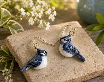 Puede incluir: Un par de pendientes colgantes con dijes de arrendajo azul pintados a mano. Cada ave muestra un vientre blanco, alas azules con detalles negros y una pequeña cresta en la cabeza. Los pájaros están unidos a ganchos de metal dorado.