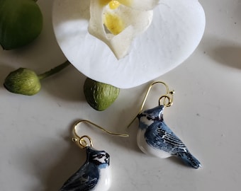 Könnte beinhalten: Ein Paar blaue und weiße Vogeleringe mit goldenen Haken. Die Vögel sind mit realistischen Details bemalt und haben einen weißen Hintergrund.