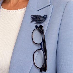 May include: A dark blue bird-shaped brooch with a textured finish, featuring a circular metal ring attached to the bottom. The brooch is pinned to a light blue blazer, and a pair of dark-rimmed eyeglasses hangs vertically from the metal ring.