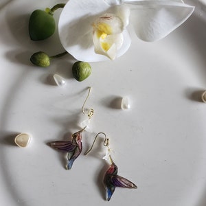 May include: A pair of gold hummingbird earrings with iridescent wings and a white flower charm. The earrings are on a white surface with white and green decorative items.