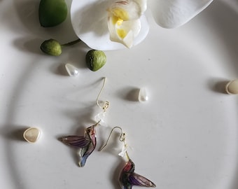 Könnte beinhalten: Ein Paar goldene Kolibri-Ohrringe mit schillernden Flügeln und einem weißen Blumen-Anhänger. Die Ohrringe liegen auf einer weißen Oberfläche mit weißen und grünen Dekorationsgegenständen.