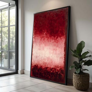 Puede incluir: Gran obra de arte abstracto con una combinación de colores rojo y blanco, enmarcada en un marco negro. La obra se apoya en una pared blanca. Una planta en maceta en una cesta tejida está cerca.