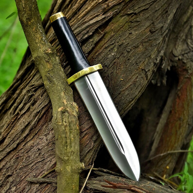 May include: A dagger with a polished silver blade and a black handle. The handle is accented with gold-colored metal at the base and top. The dagger is leaning against a textured tree trunk, showcasing its sharp design.