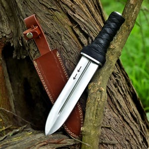 May include: A silver dagger with a black wrapped handle and a brown leather sheath. The dagger has two arrows etched on the blade. The sheath is brown with decorative stitching and a belt loop.