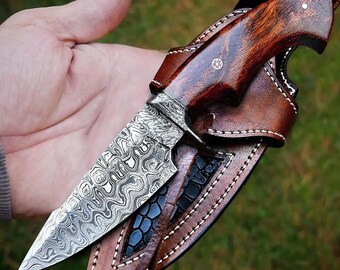 May include: A handcrafted knife with a patterned steel blade and a wooden handle. The knife is in a brown leather sheath with a decorative stitching detail.
