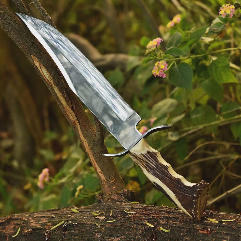 May include: A large, polished steel knife with a wide blade and a handle made of brown and cream-colored antler. The knife rests on a tree branch, with a blurred green and floral background.