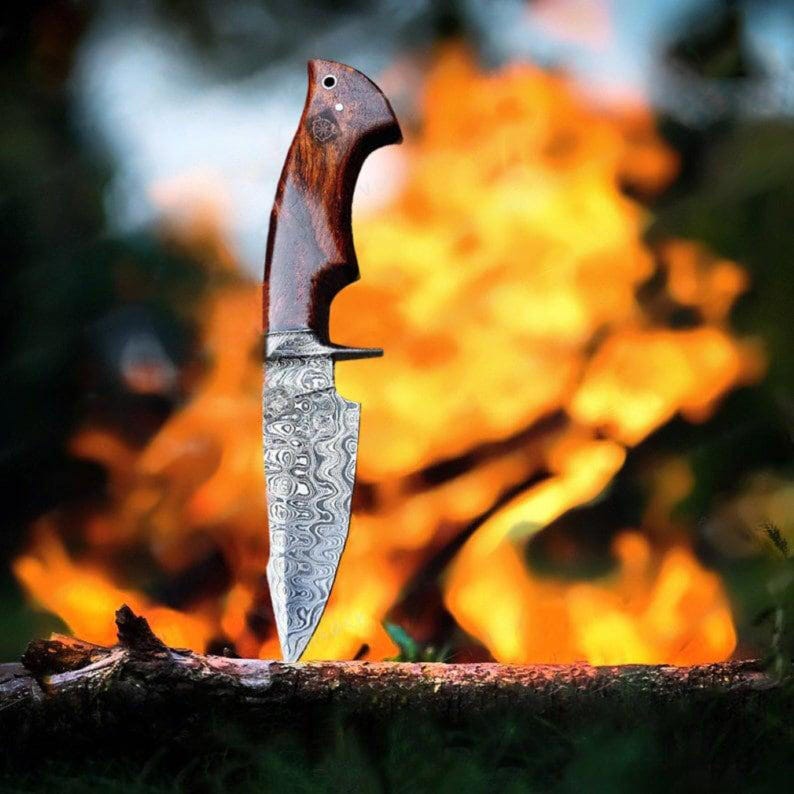 May include: A Damascus steel knife with a dark wood handle. The blade has a unique swirling pattern. The knife is resting on a log with a blurred background of flames. The knife is a hunting or camping tool.