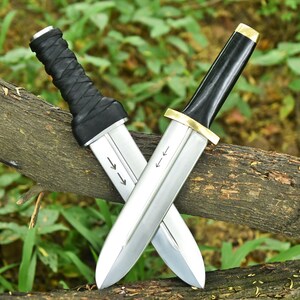 May include: Two crossed daggers with silver blades and black handles. One handle is wrapped in black leather, the other is smooth with a gold accent. The blades have etched arrows. The daggers rest on a brown branch against a green background.