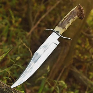 May include: A large, polished steel knife with a curved blade and a handle made of light brown and dark brown material. The knife has a guard and is set against a blurred background of green foliage and tree branches.