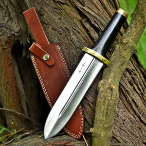 May include: A polished steel dagger with a black handle and brass accents, resting in a brown leather sheath. The blade has two etched arrows. The sheath is brown with decorative stitching and a leather strap.