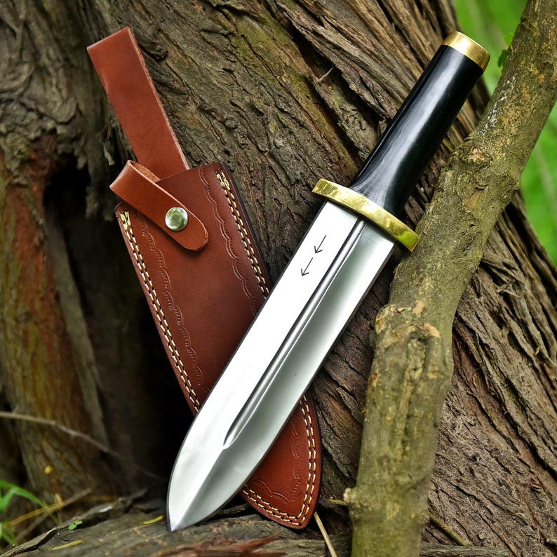 May include: A polished steel dagger with a black handle and brass accents, resting in a brown leather sheath. The blade has two etched arrows. The sheath is brown with decorative stitching and a leather strap.