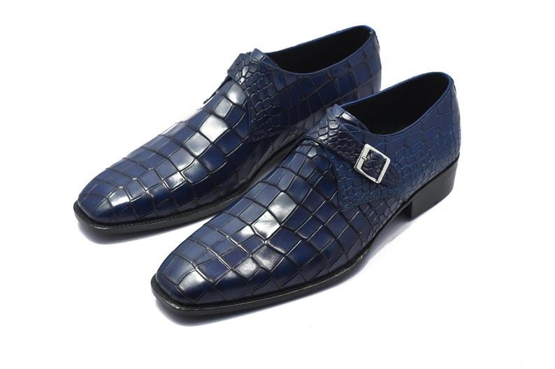 May include: A pair of navy blue leather dress shoes with a square toe and a textured, crocodile-like pattern. Each shoe features a silver buckle closure and a black sole. The shoes are displayed on a white background.