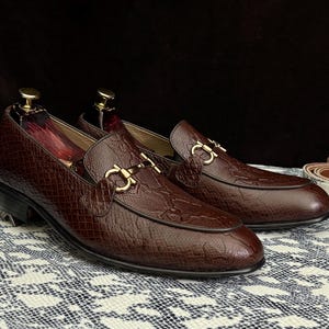 Peut inclure: Une paire de mocassins en cuir marron avec des boucles dorées. Les mocassins ont un motif texturé et gaufré et sont posés sur une surface à motifs blancs et noirs.