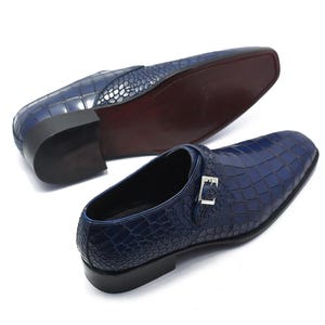 May include: Two navy blue leather dress shoes with a crocodile skin texture. One shoe is upright, and the other is upside down, revealing a dark brown sole. Each shoe has a silver buckle.