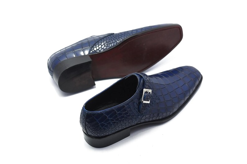 May include: Two navy blue leather dress shoes with a crocodile skin texture. One shoe is upright, and the other is upside down, revealing a dark brown sole. Each shoe has a silver buckle.