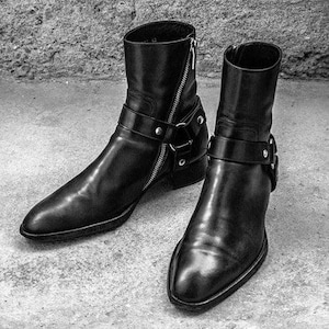 Könnte beinhalten: Ein Paar schwarze Leder-Stiefeletten mit seitlichem Reißverschluss und einem Schnallenriemen. Die Stiefeletten haben eine spitze Zehenpartie und einen niedrigen Absatz.