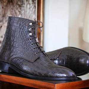 May include: A pair of brown leather ankle boots with a textured, crocodile-like pattern. The boots have a lace-up design with silver-toned eyelets and a small, square heel. The boots are displayed on a wooden surface.