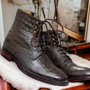 May include: A pair of dark brown leather boots with a crocodile skin texture. The boots have laces, metal eyelets, and a zipper on the side. The boots are resting on a white, fluffy surface.