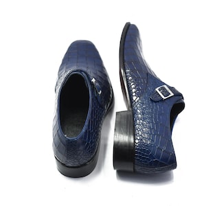 May include: A pair of navy blue leather dress shoes with a crocodile skin texture. The shoes have a square toe and a buckle closure. The soles are black.