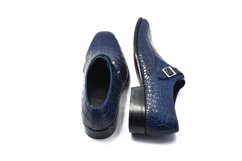 May include: A pair of navy blue leather dress shoes with a crocodile skin texture. The shoes have a square toe and a buckle closure. The soles are black.
