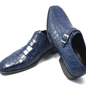 May include: A pair of blue leather dress shoes with a crocodile skin pattern. The shoes have a square toe and a silver buckle closure. The shoes are on a white background.