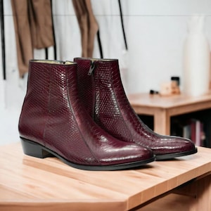 Op de afbeelding: Een paar bordeauxkleurige enkellaarsjes met een textuurpatroon dat op slangenleer lijkt. De laarzen hebben een spitse neus, een lage hak en een ritssluiting. Ze staan op een licht houten oppervlak.