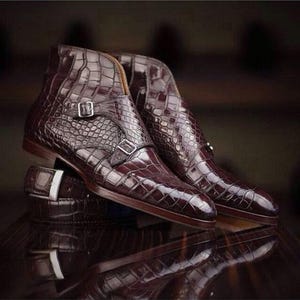 May include: Burgundy leather ankle boots with a crocodile skin effect. The boots feature two silver buckles and a stacked heel. A matching belt with a silver buckle is also visible.