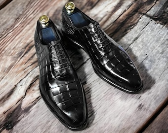 May include: A pair of black leather dress shoes with a textured, checkered pattern. The shoes have laces and are displayed on a wooden surface. Shoe trees are inserted in the shoes.