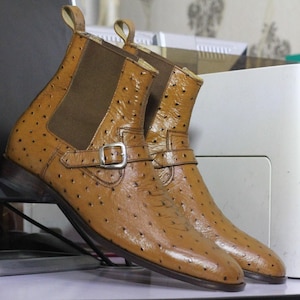 May include: A pair of tan leather ankle boots with a textured surface and a buckle detail. The boots have elastic side panels and pull tabs. The footwear is displayed on a light-colored surface.