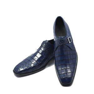 May include: A pair of navy blue leather dress shoes with a textured, crocodile-like pattern. Each shoe features a silver-colored buckle closure. The shoes have a square toe and a low heel.
