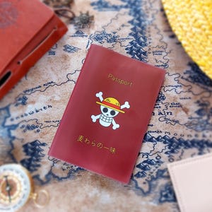 May include: A red passport cover with the word "Passport" printed on it, featuring a cartoon skull and crossbones with a straw hat. The cover also has Japanese text. Other travel accessories are in the background.