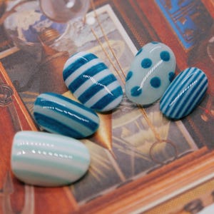 Peut inclure: Un ensemble de cinq faux ongles ovales arborant divers motifs bleus et blancs. Les designs incluent des rayures horizontales de largeurs variées, ainsi qu'une base bleu clair avec des pois bleu foncé, le tout avec une finition brillante.