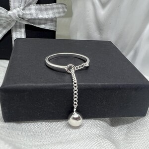 Könnte beinhalten: Ein Ring aus poliertem Silber mit einer zarten Kette, die am Ringband befestigt ist und in einer kleinen, glatten, kugelförmigen Silberperle endet. Der Ring hat ein schlichtes, massives Banddesign mit einer hellen, reflektierenden Oberfläche.