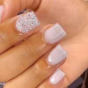 May include: Close-up of a hand with short, square-shaped nails painted a light, milky white. One nail features a cluster of small, clear rhinestones.
