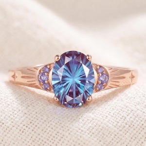 Op de afbeelding: Een roségouden ring met een grote ovaal geslepen blauw en paars van kleur veranderende middensteen. De band is versierd met kleine ronde paarse zijstenen en gegraveerde zonnestraalpatronen aan beide kanten van de zetting.