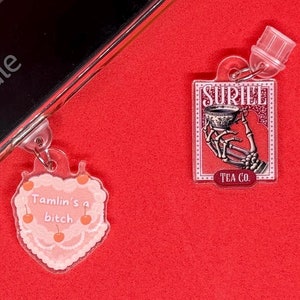 Könnte beinhalten: Zwei Acryl-Anhänger mit Schlüsselanhängern auf rotem Hintergrund. Ein Anhänger ist herzförmig mit einem Kuchen-Design und dem Text "Tamlin's a bitch". Der andere ist rechteckig mit einer Skeletthand, die eine Tasse hält, und dem Text "Suriel Tea Co."